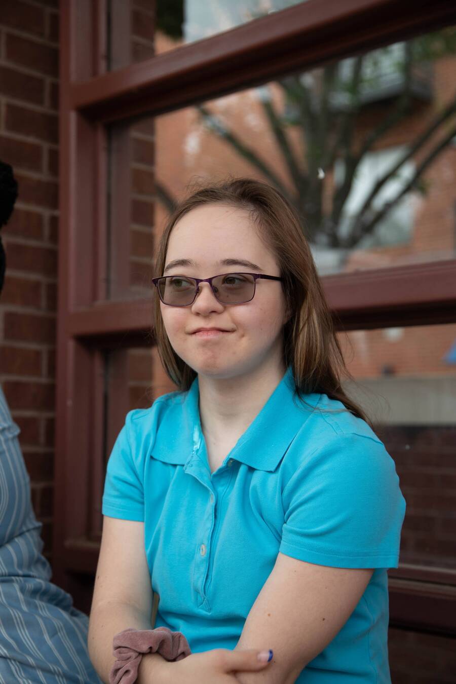 A smiling young woman in a teal shirt sits outside a brick building.