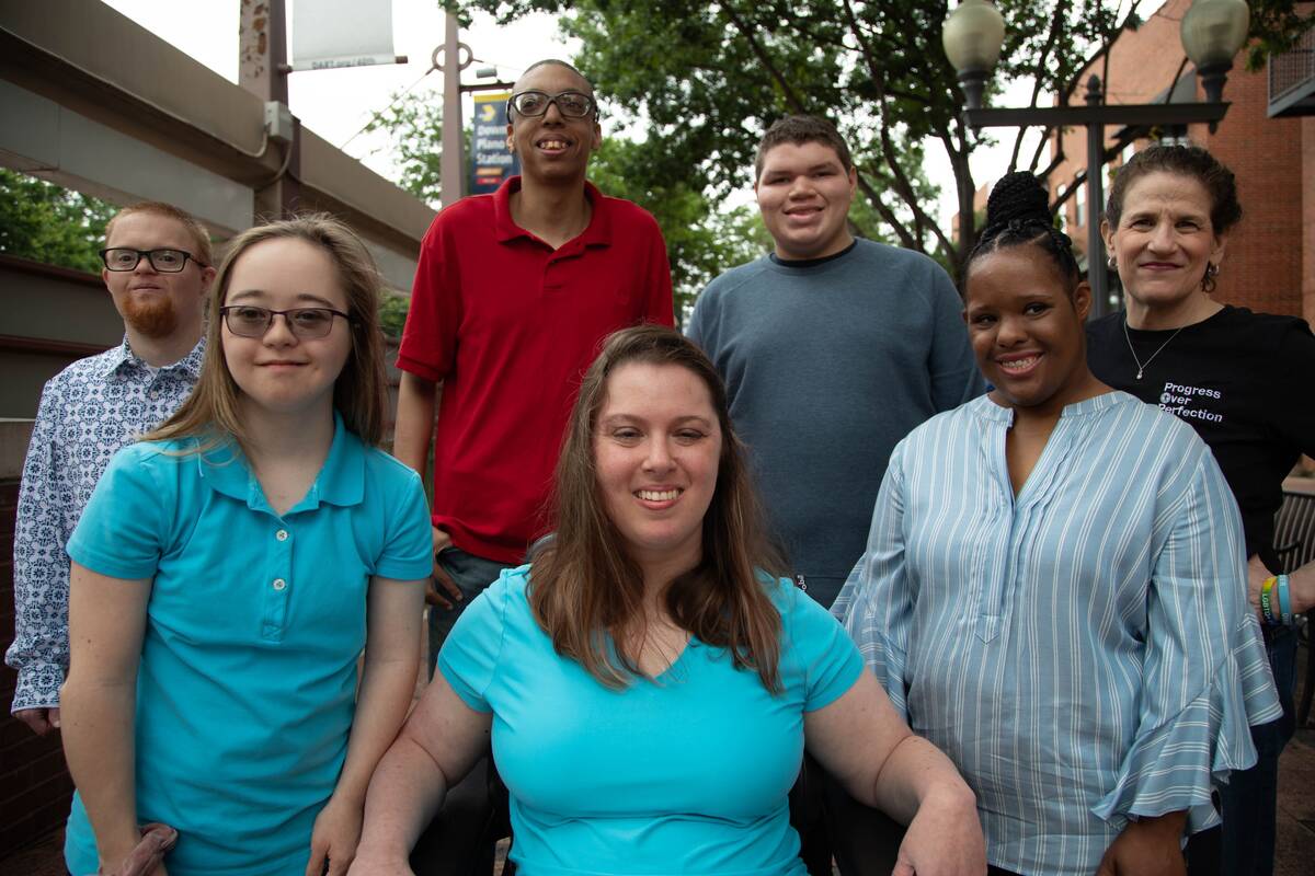 A group of friends with and without disabilities smile together on a city sidewalk.