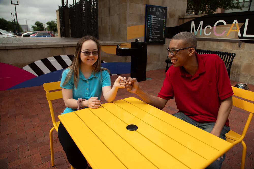 Two friends fist bump across a yellow table on a plaza.