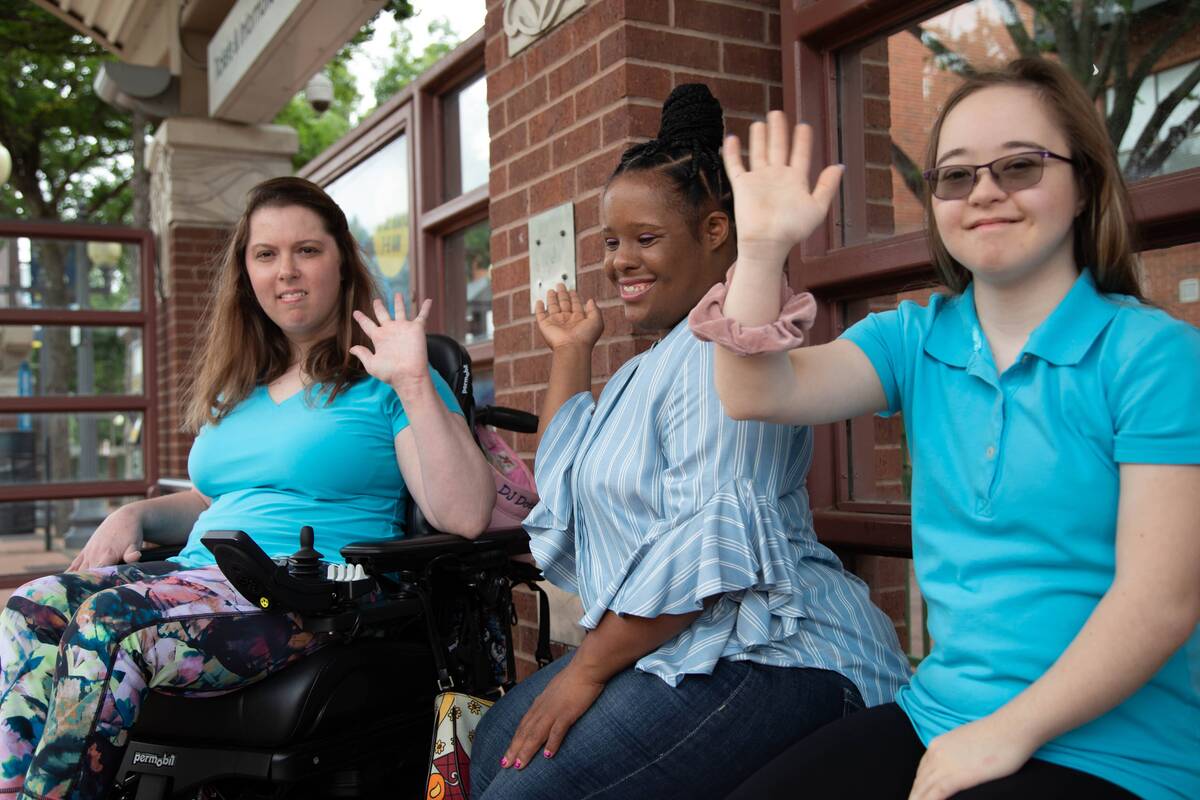 Three friends, one using a power wheelchair, wave and smile outside a train station.